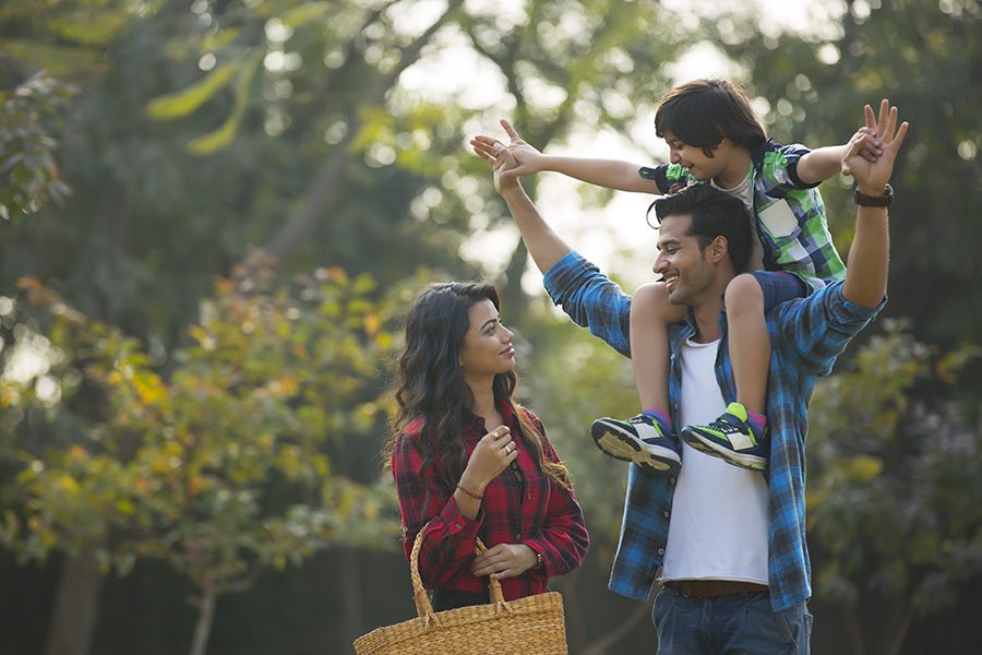 About Our Agency - Happy Young Couple Standing in a Garden With the Man Carrying Their Son on His Shoulders and the Woman Carrying a Basket