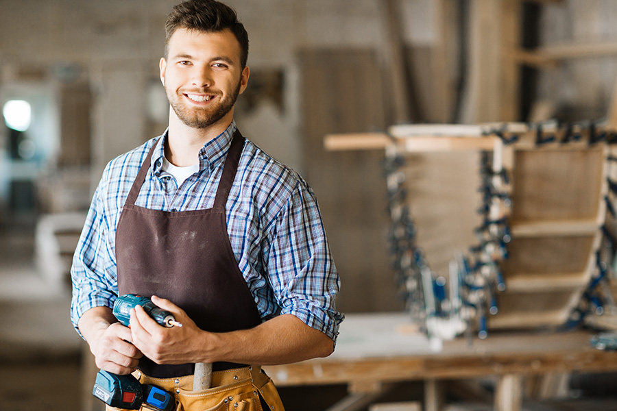 Specialized Business Insurance - Portrait of Smiling Bearded Craftsman With Electric Drill in Hands Standing in Spacious Workshop
